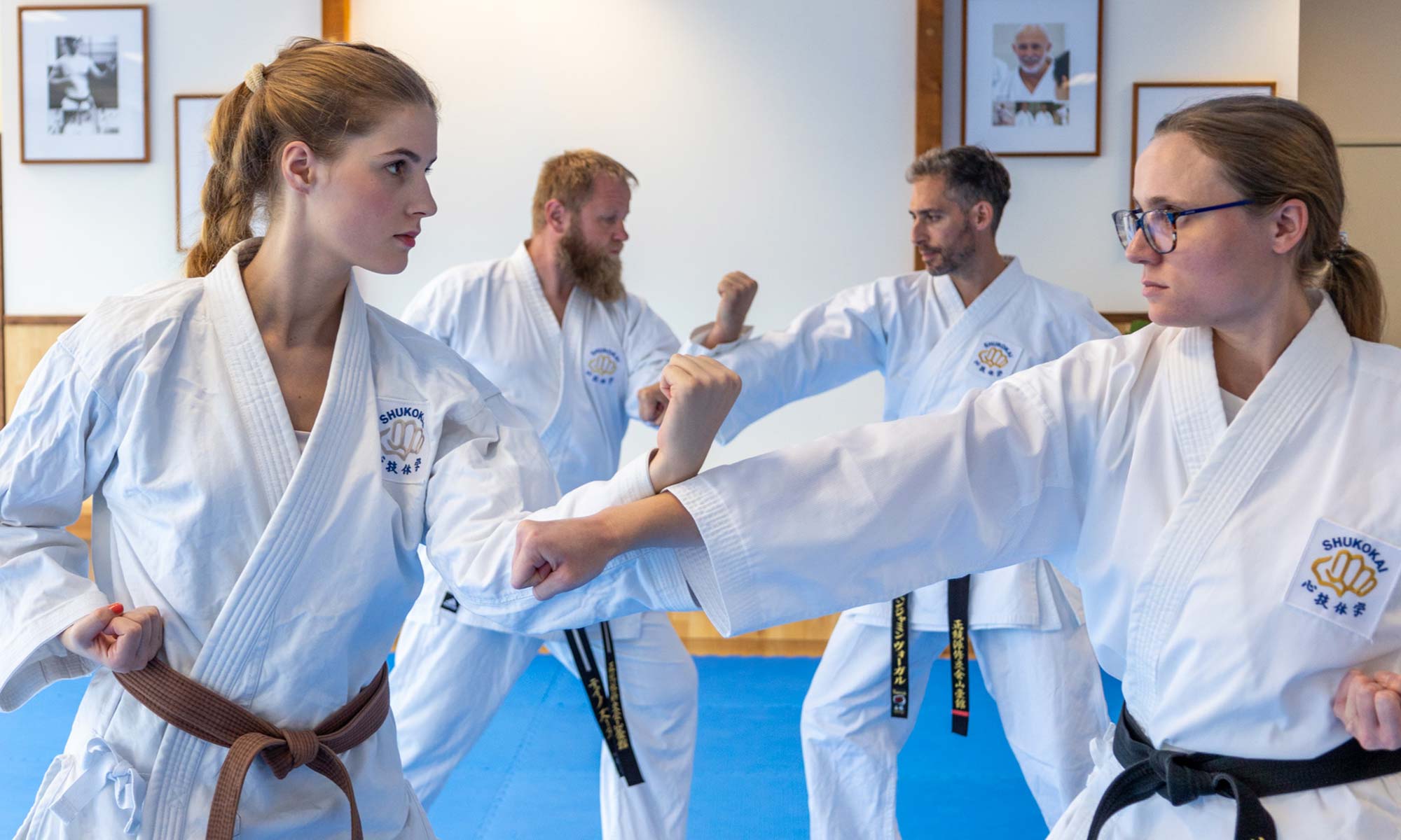 Das Bild zeigt vier Personen in Karateuniformen (Gi), die in einem Dojo Kampfsporttechniken üben. Eine Person trägt einen braunen Gürtel und die anderen drei tragen schwarze Gürtel, was auf ihren Rang hinweist. Sie nehmen eine Haltung oder Bewegung ein, bei der sie sich mit ausgestreckten Fäusten gegenüberstehen. Im Hintergrund sind gerahmte Fotos an der Wand zu sehen, die die traditionelle Kampfsportkulisse unterstreichen. Das Logo auf ihren Uniformen lautet „Shukokai“, eine Stilrichtung des Karate.