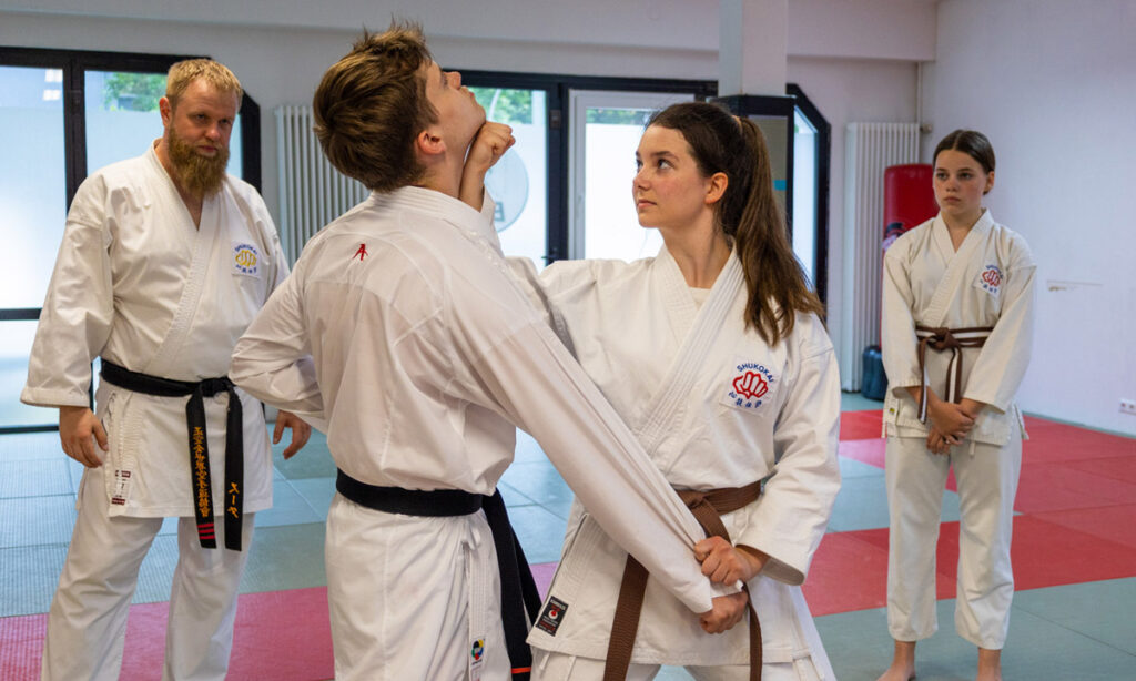 Eine Gruppe von Menschen übt Karate in einem Dojo. Ein Ausbilder mit schwarzem Gürtel beobachtet, wie eine junge Frau mit weißem Karateanzug und braunem Gürtel eine Technik an einem jungen Mann mit weißem Karateanzug und schwarzem Gürtel ausführt. Eine andere junge Frau mit einem braunen Gürtel schaut im Hintergrund aufmerksam zu. Der Schauplatz ist ein Dojo mit Matten auf dem Boden.