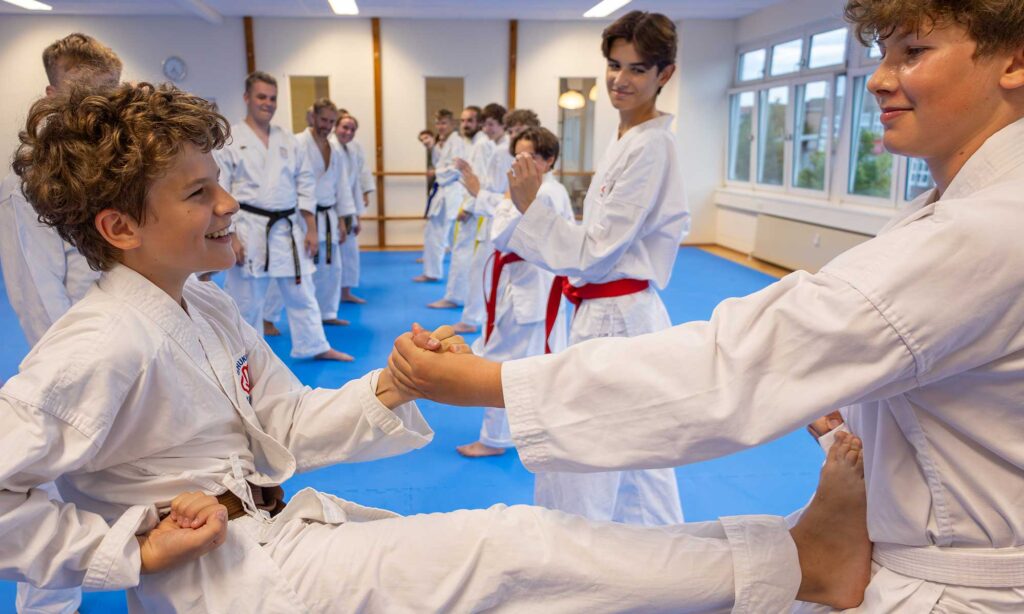 Zwei jugendliche Karateschüler üben zusammen in einem Dojo. Sie lächeln, während der eine einen kontrollierten Tritt ausführt, während der andere seinen Fuß festhält. Im Hintergrund sind andere Schüler und Ausbilder mit schwarzen Gürteln zu sehen, die das Training beobachten und daran teilnehmen.