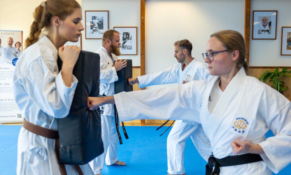 Das Bild zeigt eine Gruppe von Kampfsportlern beim Karatetraining. Es sind zwei Männer und zwei Frauen, alle in weißen Karateuniformen (Gi) mit Gürteln, die ihre Ränge kennzeichnen. Die Teilnehmer üben Schläge, indem sie auf gepolsterte Schilde schlagen, die von ihren Partnern gehalten werden. Die Trainingsumgebung scheint ein Dojo zu sein, mit Holzwänden und gerahmten Kampfsportfotos im Hintergrund.