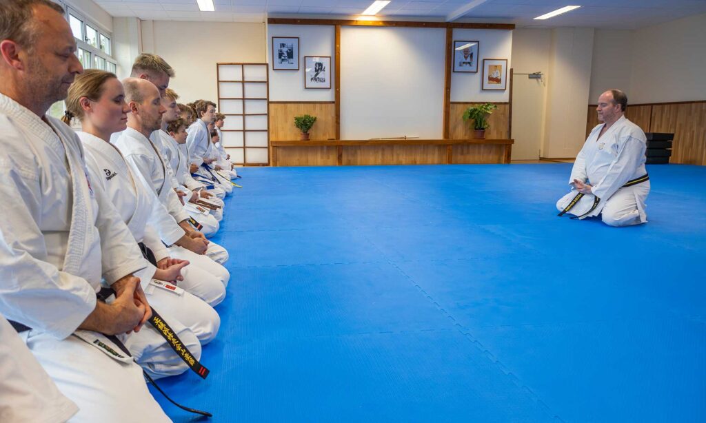 Eine Gruppe von Karate-Schülern in weißen Uniformen sitzt in kniender Position auf einer blauen Matte in einem Dojo. Die Atmosphäre ist ruhig und meditativ, während die Schüler und der Lehrer einen Moment der Konzentration praktizieren. Das Dojo hat eine Holzverkleidung, gerahmte Kampfsportfotos und eine minimale Dekoration im Hintergrund.