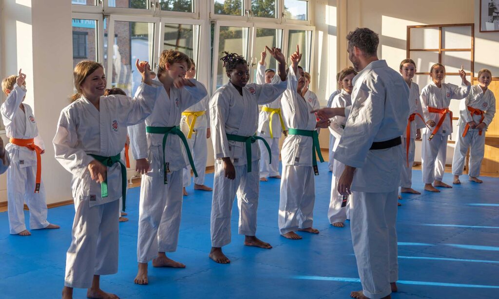 Eine Gruppe von Kindern in Karate-Uniformen und mit verschiedenfarbigen Gürteln nimmt an einer Unterrichtsaktivität teil und hebt die Hand, um eine Frage zu beantworten oder mitzumachen. Der Ausbilder, der einen schwarzen Gürtel trägt, interagiert mit den Schülern in einem hellen Dojo mit großen Fenstern.