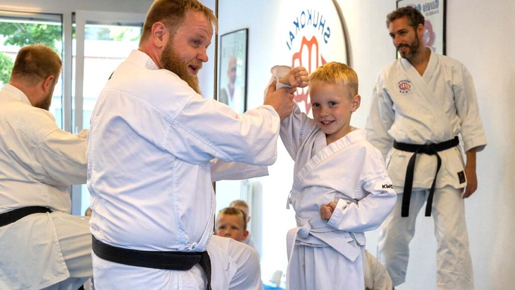 Ein Kampfsportlehrer in einem weißen Karate-Gi hilft einem kleinen Jungen, ebenfalls in einem weißen Karate-Gi, bei einer Technik. Beide lächeln, und der Junge sieht begeistert aus. Im Hintergrund sind andere Schüler und Ausbilder zu sehen, die das Training beobachten. Die Umgebung ist ein Dojo.