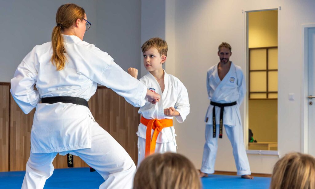 Ein Kind übt Karate mit einem orangefarbenen Gürtel und führt eine Blocktechnik unter Anleitung eines erwachsenen Ausbilders mit schwarzem Gürtel aus, während ein anderer Ausbilder im Hintergrund zuschaut. Andere Kinder sitzen und beobachten den Unterricht.